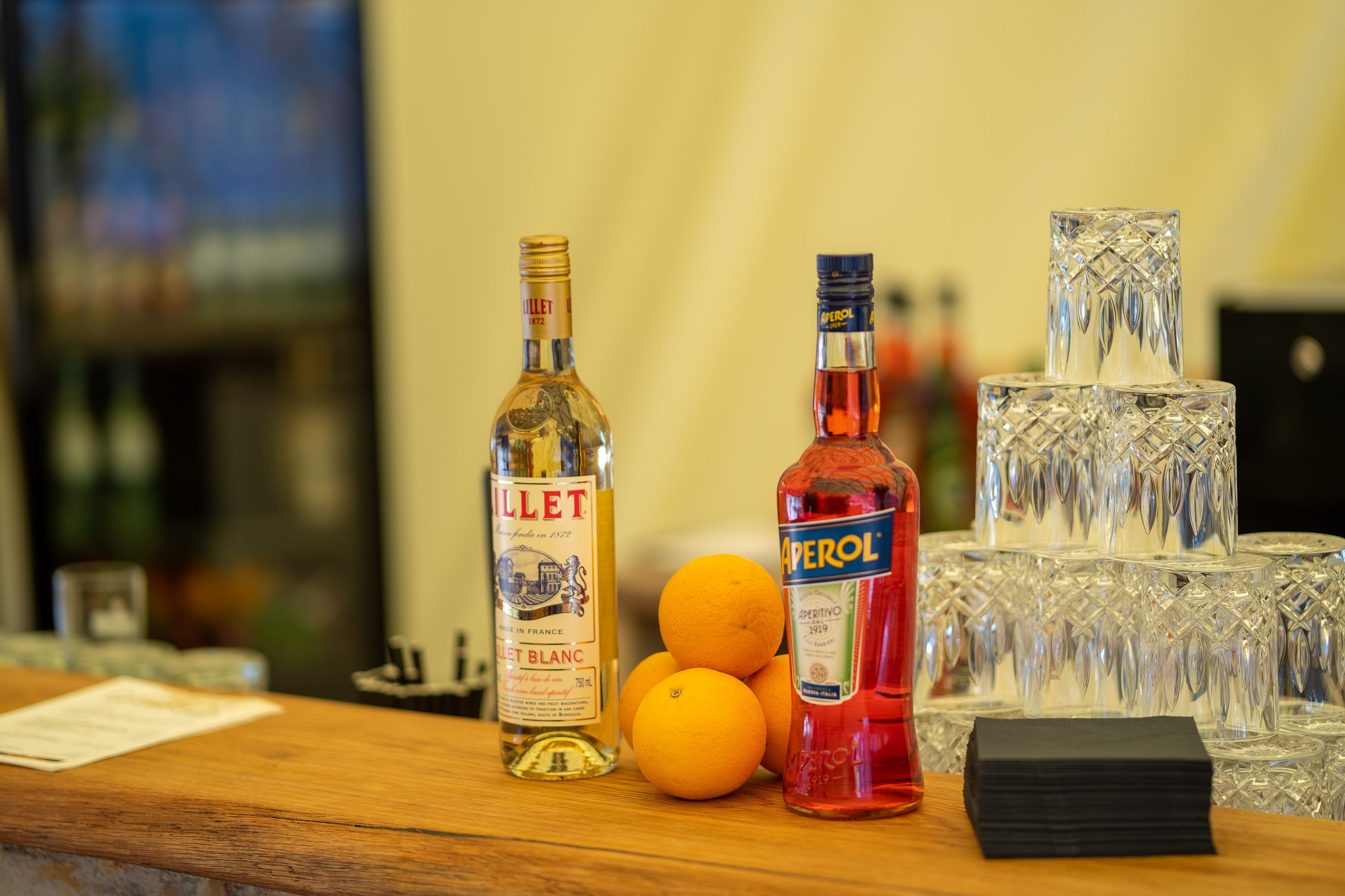 A bar area with bottles of liquor, stacked glasses, and oranges on a wooden counter.