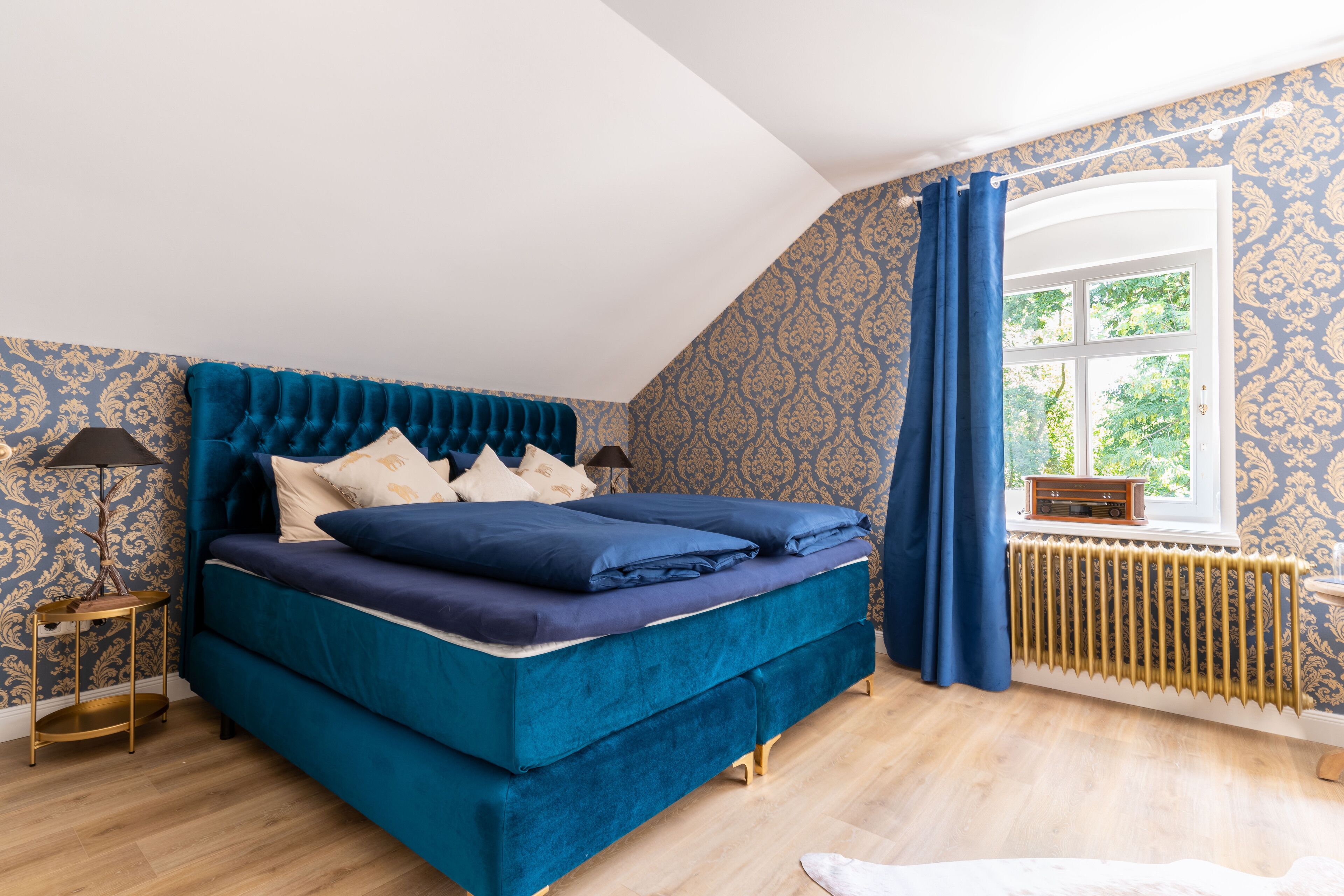 A bedroom with a large bed, a window with curtains, a radiator, and patterned wallpaper.