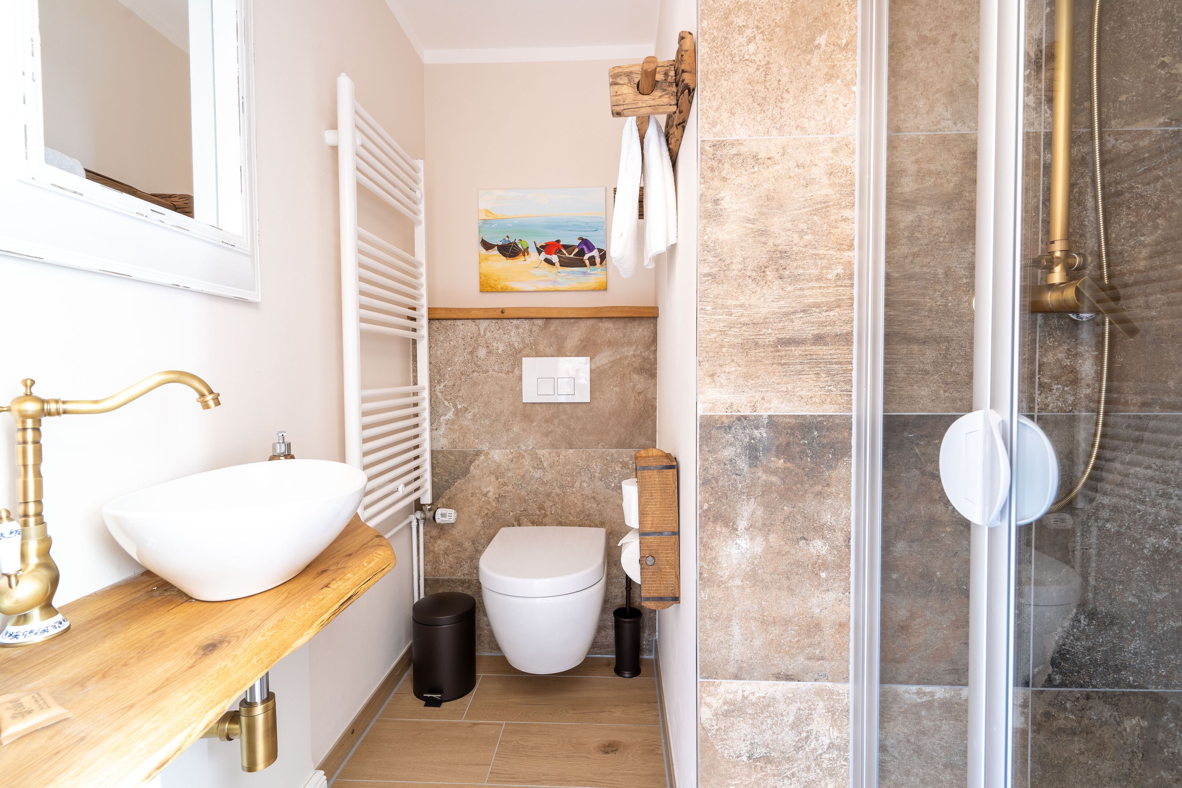 A bathroom with a white sink, toilet, and shower. There is a wooden shelf above the toilet and a painting on the wall.