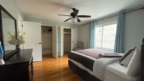 Ein Schlafzimmer mit Bett, Deckenventilator, Fenster mit Vorhängen und Holzboden.
