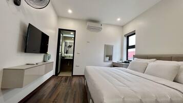 A modern hotel room with a large bed, a TV mounted on the wall, a desk with a chair, and a window providing natural light.