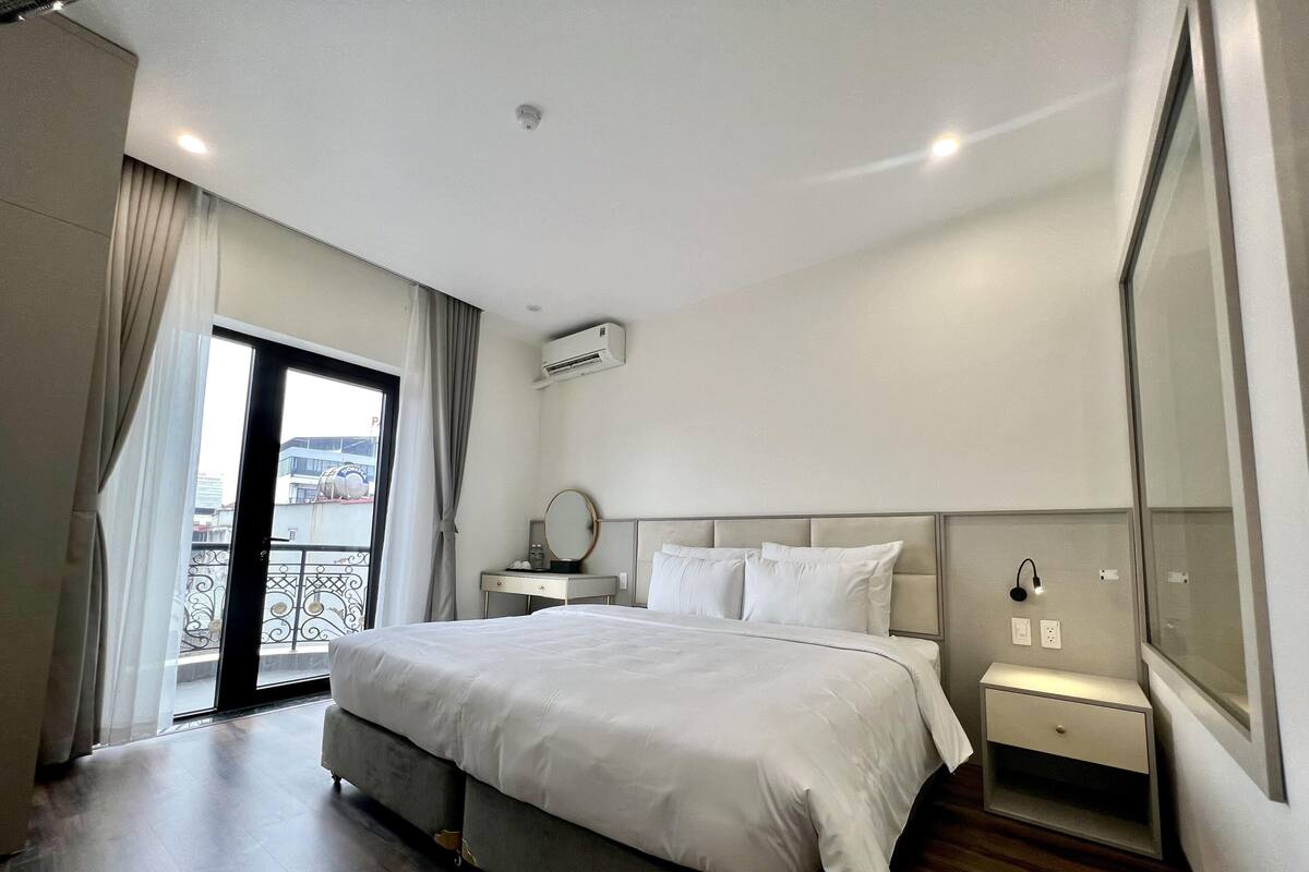 Una habitación de hotel moderna con una cama grande, una mesita de noche con lámpara, un equipo de aire acondicionado y vistas de la ciudad a través de las puertas del balcón.