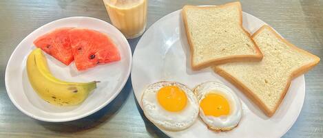 A plate with two fried eggs, slices of bread, a banana, and a slice of watermelon on a table.