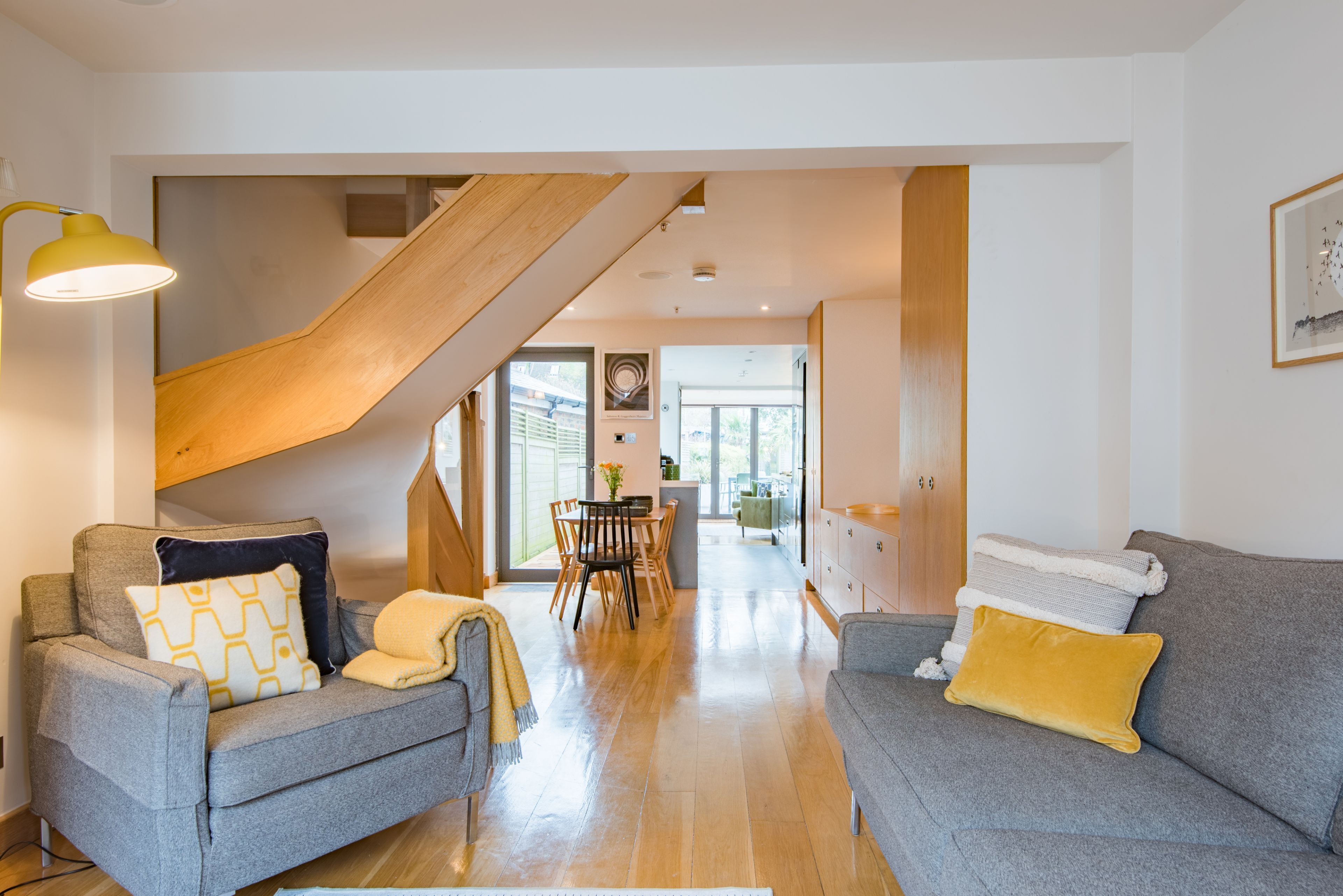 A modern living room with two grey sofas, a wooden staircase, and a dining area in the background.