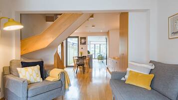 A modern living room with two grey sofas, a wooden staircase, and a dining area in the background.