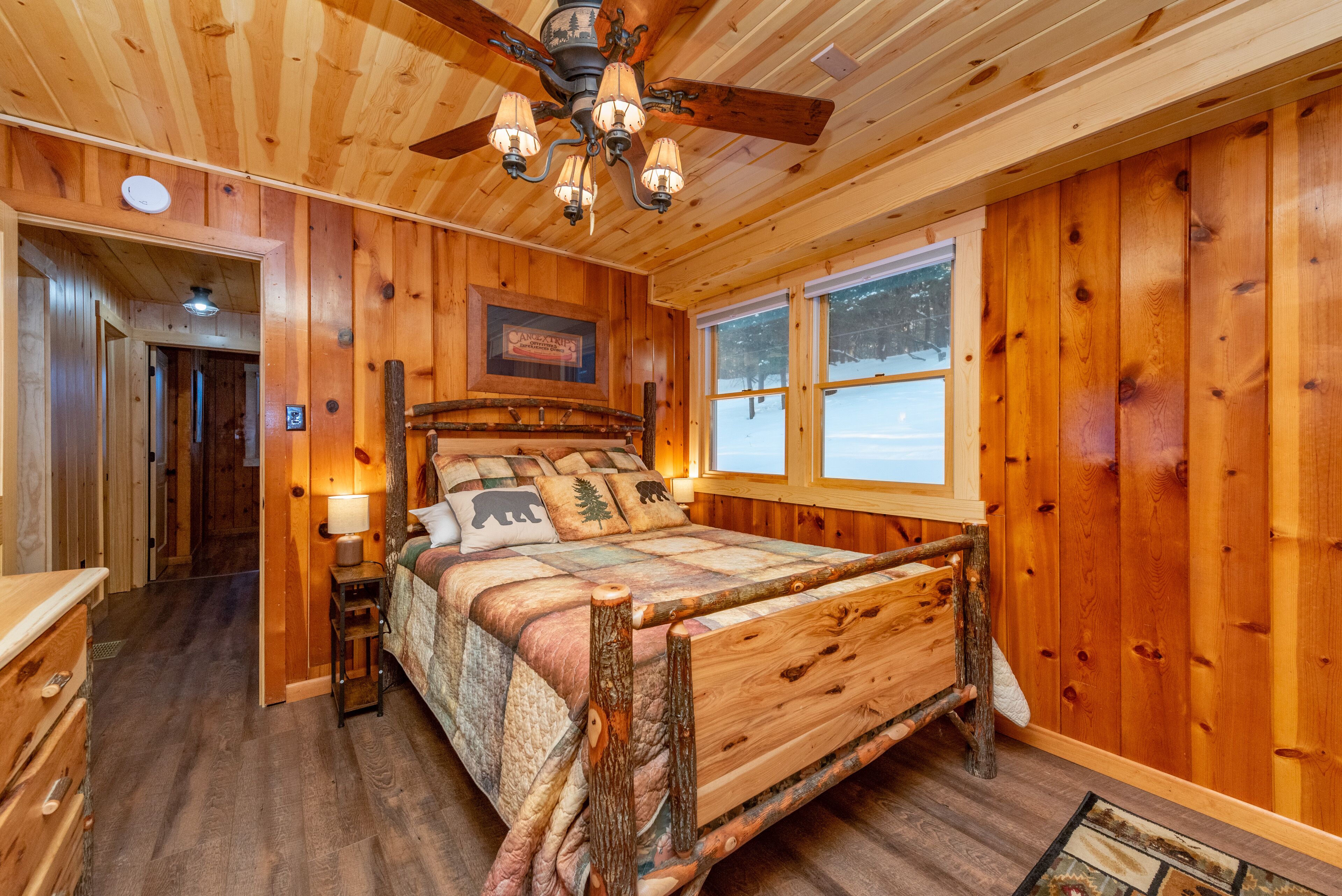 A wooden cabin bedroom with a bed, a ceiling fan, a window with a view of the outside, and a wooden dresser.