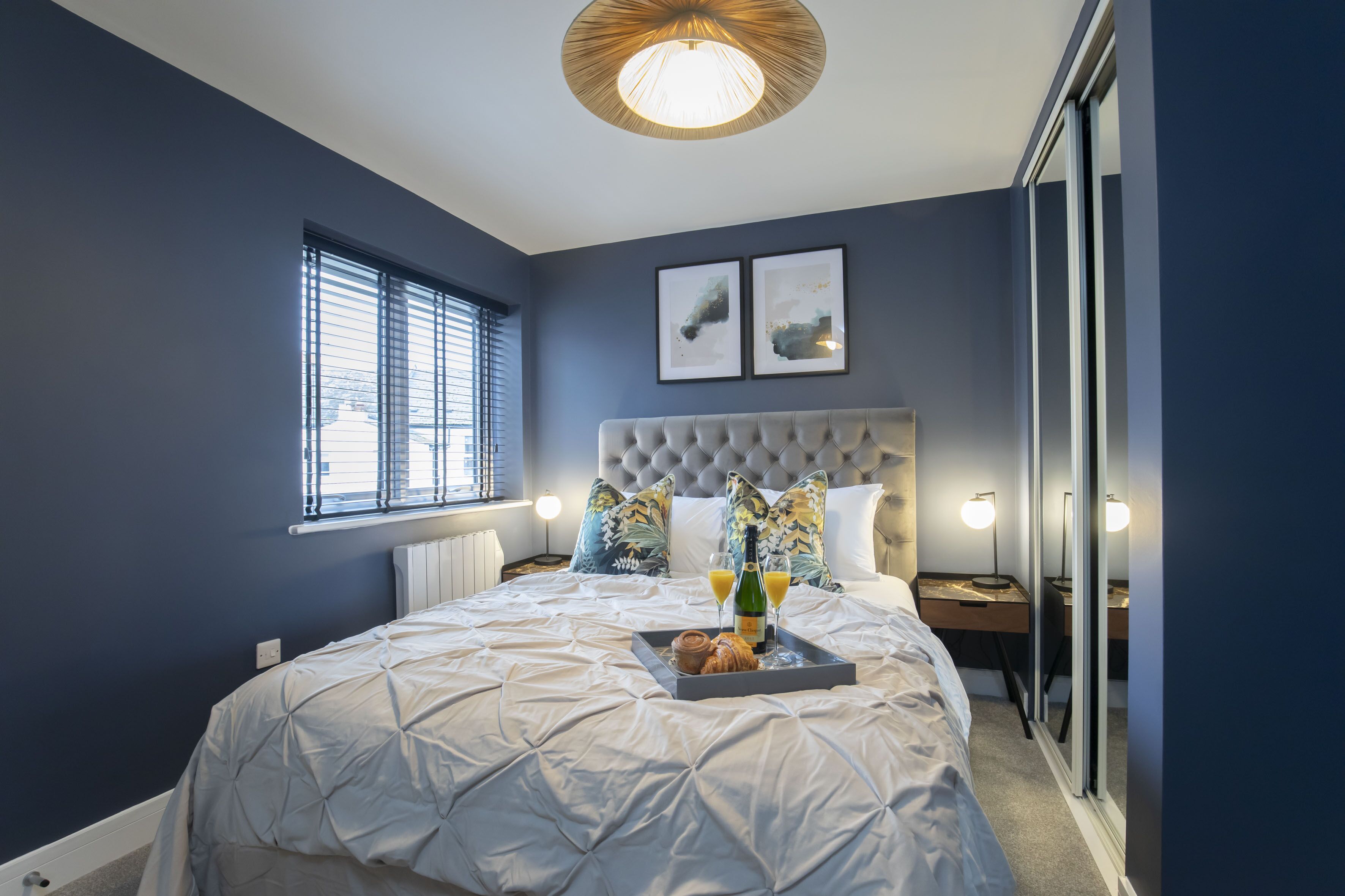 A bedroom with a large bed, a bedside table, a nightstand, a window with blinds, and two framed pictures on the wall.
