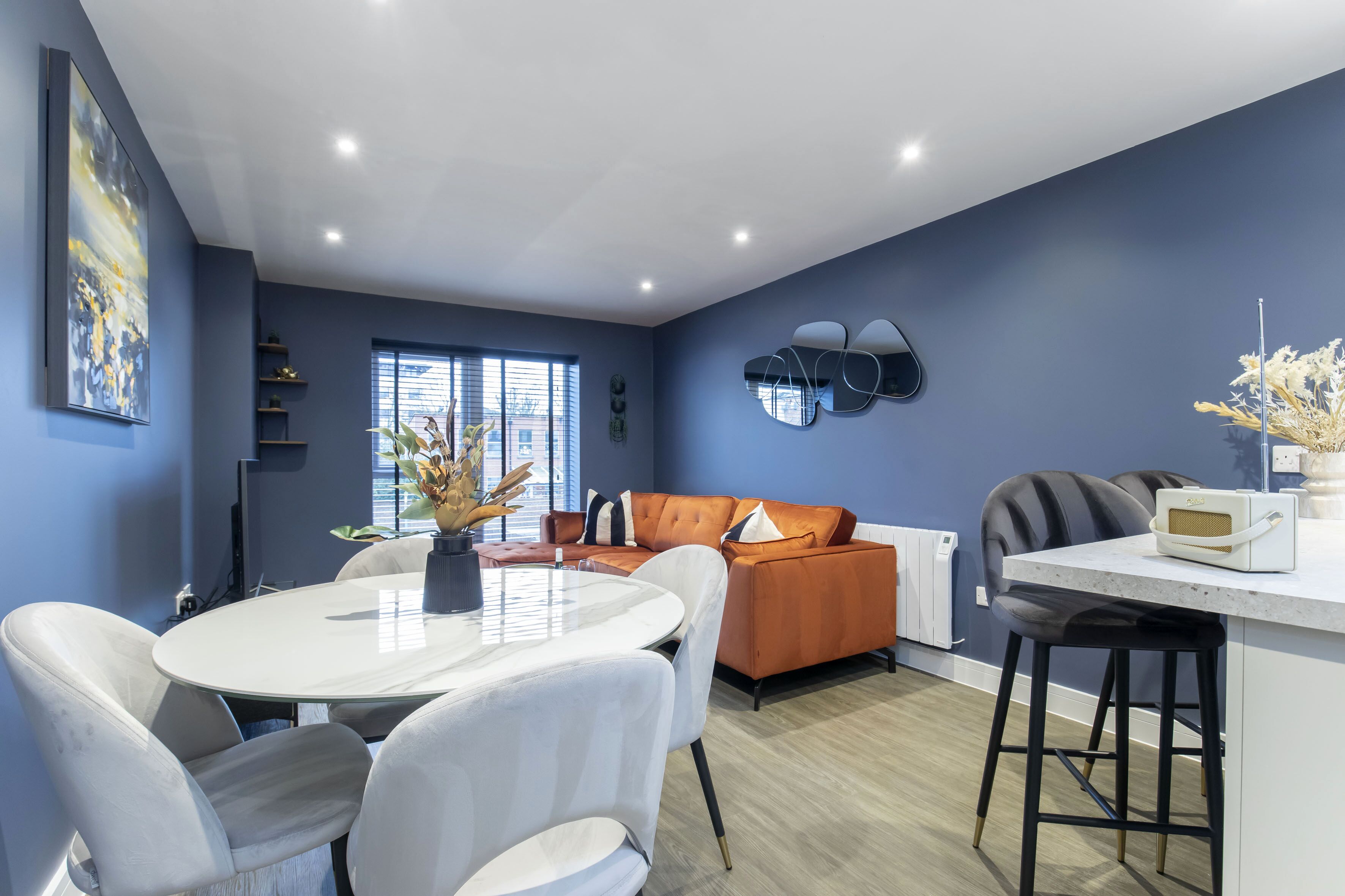 A modern living room with a blue accent wall, a white round dining table, and a sofa with orange cushions.