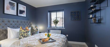 Une chambre à coucher avec une tête de lit capitonnée, un grand lit avec une literie blanche, un plateau avec des croissants et du jus, et une fenêtre avec des stores.