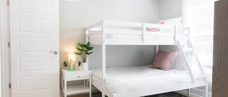 En køjeseng med skrivebord og en plante, en hvid dør og et vindue med persienner.