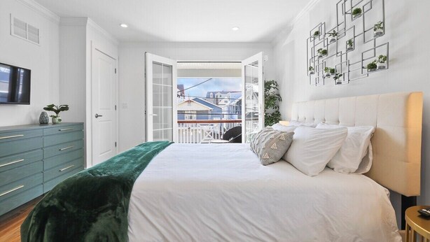 A bedroom with a large bed, a green velvet throw, a white headboard, a TV on the left, and a balcony with a view of buildings.