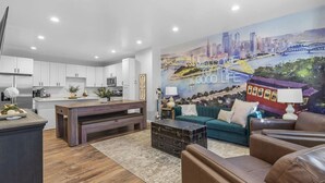 A modern kitchen with white cabinets, a wooden dining table, and a cityscape mural on the wall.