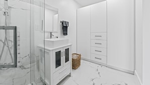 A modern bathroom with a glass-enclosed shower, white marble flooring, a white vanity with a mirror, and a large wardrobe with multiple drawers.