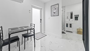 A modern, minimalist room with a dining table, chairs, a white door, a bathroom with a sink and mirror, and a framed artwork on the wall.