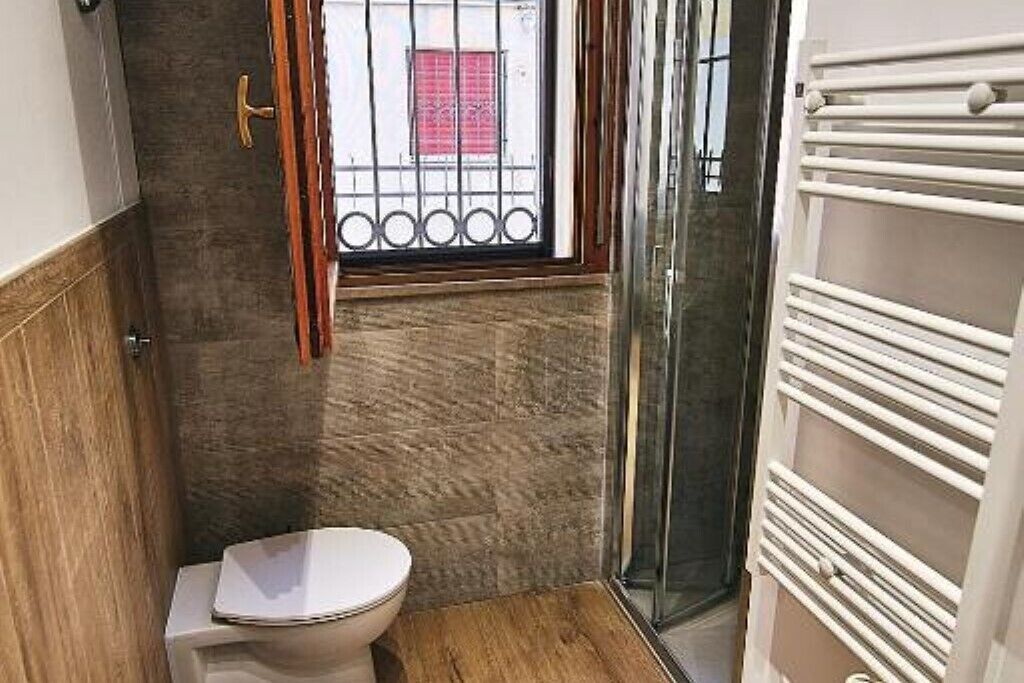 A compact bathroom with a toilet, a window with metal bars, and a glass shower enclosure.
