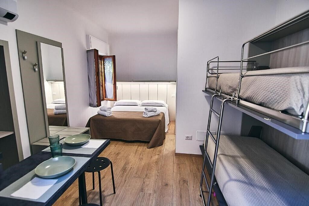 A compact hotel room with a bunk bed, a single bed, a bedside table with a mirror, and a small desk with a chair.