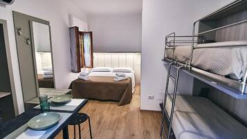 A compact hotel room with a bunk bed, a single bed, a bedside table with a mirror, and a small desk with a chair.
