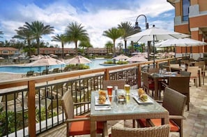 Zona de comedor junto a la piscina con mesas y sillas, con vistas a una piscina y palmeras.