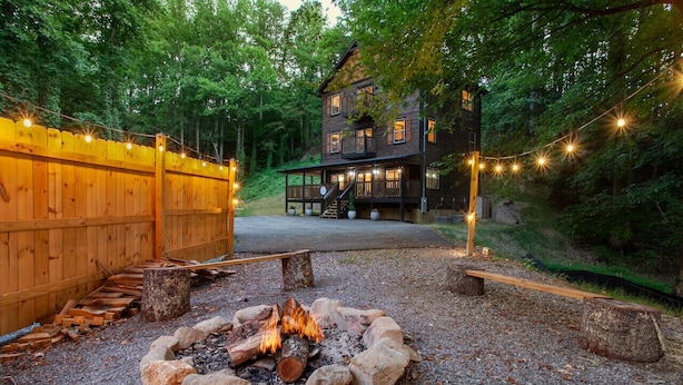 Une cabane rustique avec un foyer à l’extérieur, entourée d’une clôture en bois et illuminée de guirlandes lumineuses.