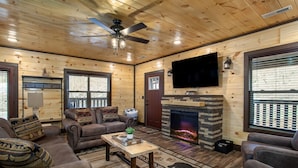 Un intérieur de chalet chaleureux avec une cheminée, des murs en bois, un ventilateur de plafond et un téléviseur à écran plat.