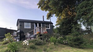 A two-story house with a dark exterior, a visible chimney, and a front porch with a flower basket. - 1 Bedroom Cabin Sleeps 2, Private Garden, Wifi (Camelford)