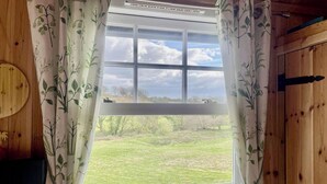 A window with a view of a grassy field and trees, framed by curtains with a leaf pattern. - 1 Bedroom Cabin Sleeps 2, Private Garden, Wifi (Camelford)
