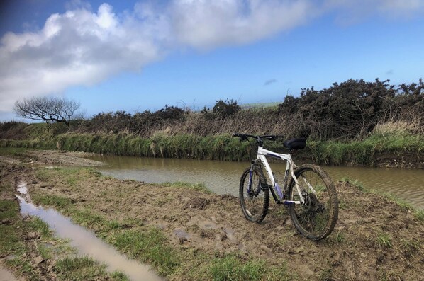 A white mountain bike parked on a muddy path beside a calm river, surrounded by natural vegetation and trees. - 1 Bedroom Cabin Sleeps 2, Private Garden, Wifi (Camelford)