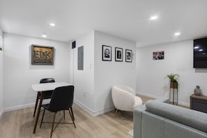 A modern living room with a dining area, featuring a white table, black chairs, a grey sofa, and framed pictures on the wall.