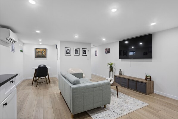 A modern living room with a grey sofa, a dining table with chairs, a TV mounted on the wall, and a wooden cabinet.