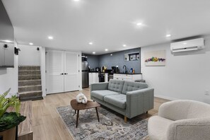 Una sala moderna con un sofá gris, un sillón blanco y una mesa de centro. Se ve una cocina al fondo con armarios blancos y una encimera negra.