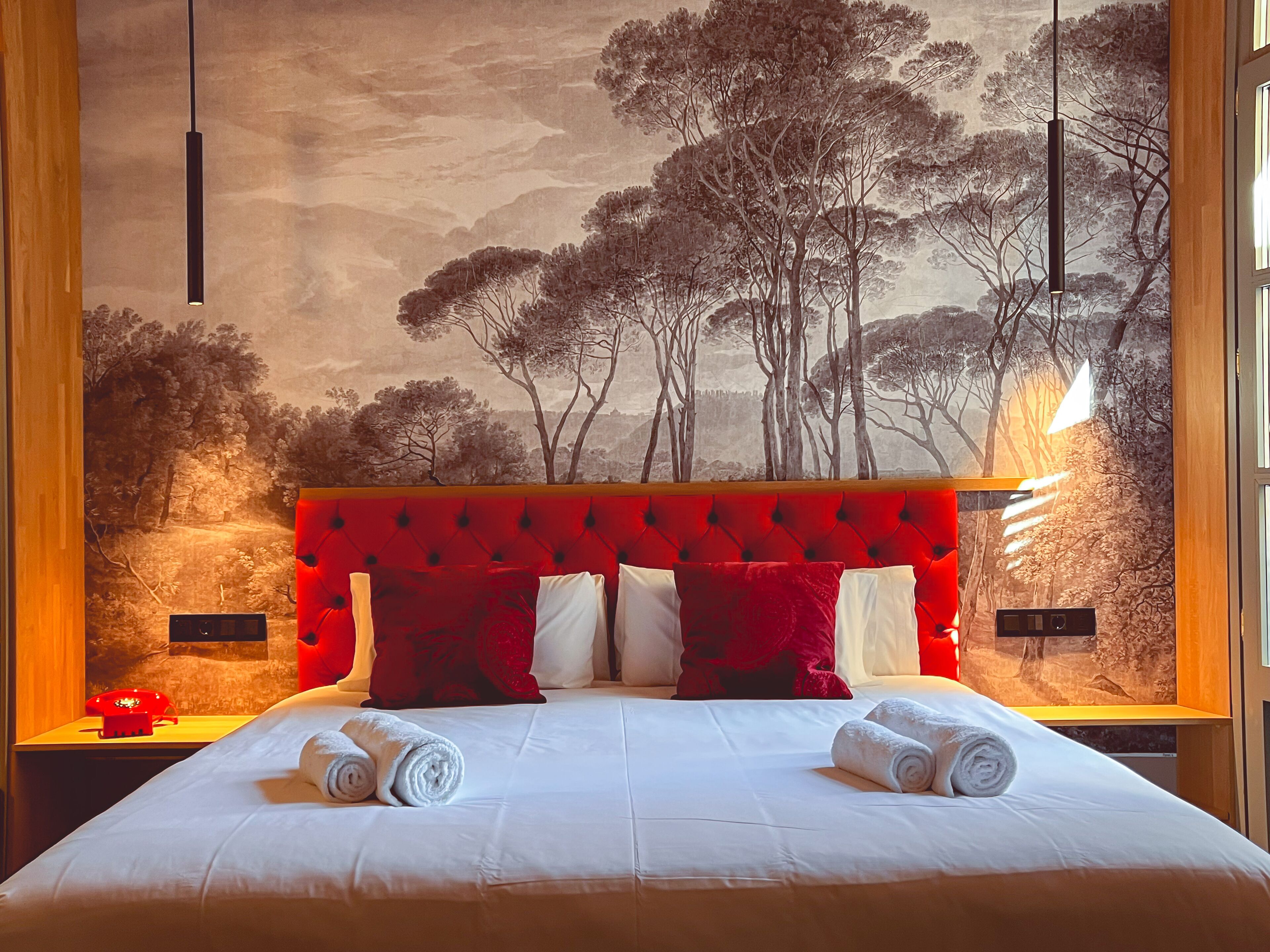 Een hotelkamer met een groot bed, rode en witte kussens en een muurschildering van bomen.