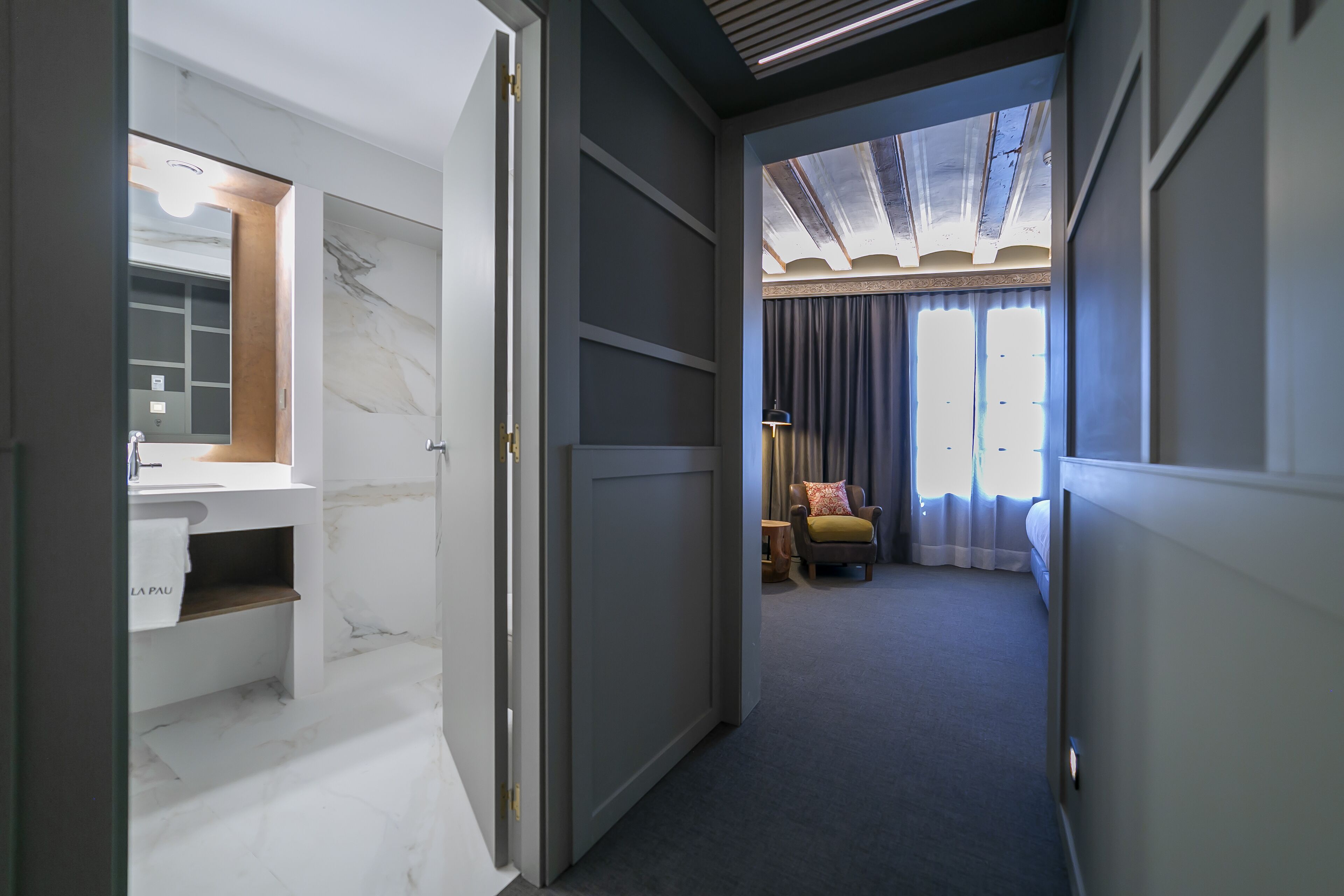 A hallway leading to a bedroom with a large window, a bedside table, and a lamp.