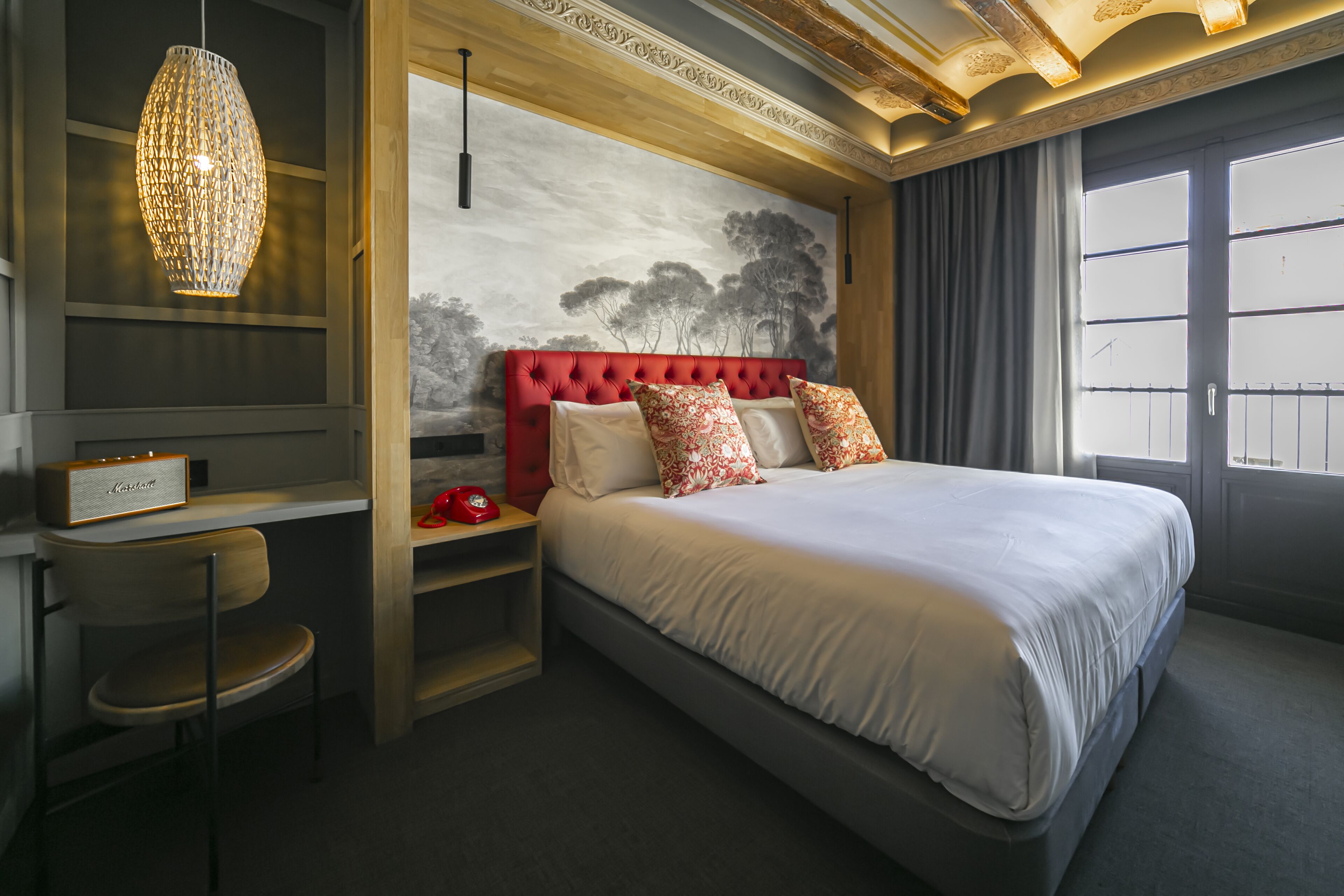Une chambre avec un grand lit, une tête de lit rembourrée de tissu rouge, un téléviseur fixé au mur, une chaise en bois et une grande fenêtre avec des rideaux.