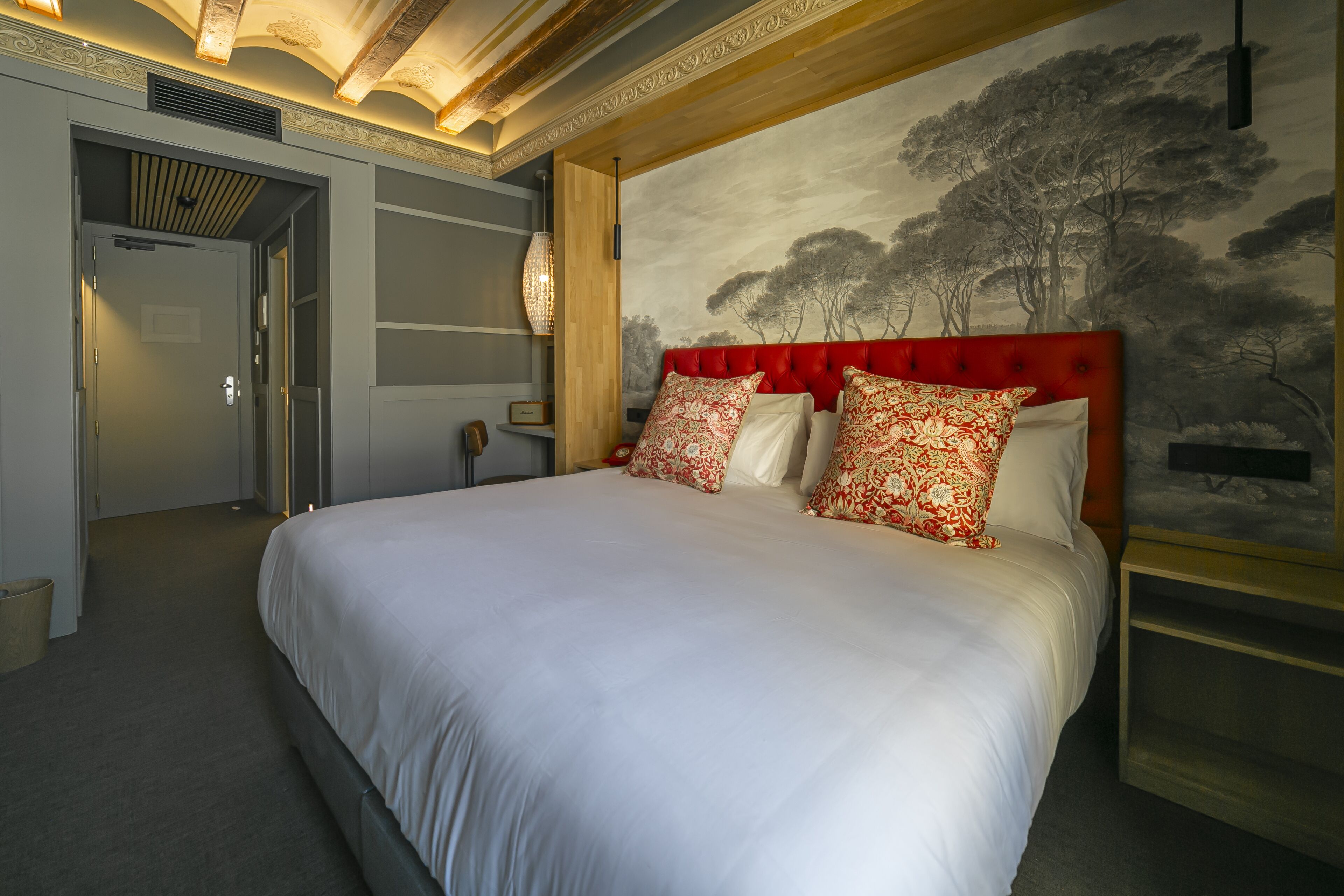 Une chambre à coucher avec un grand lit, une tête de lit rouge et une fresque représentant des arbres sur le mur.