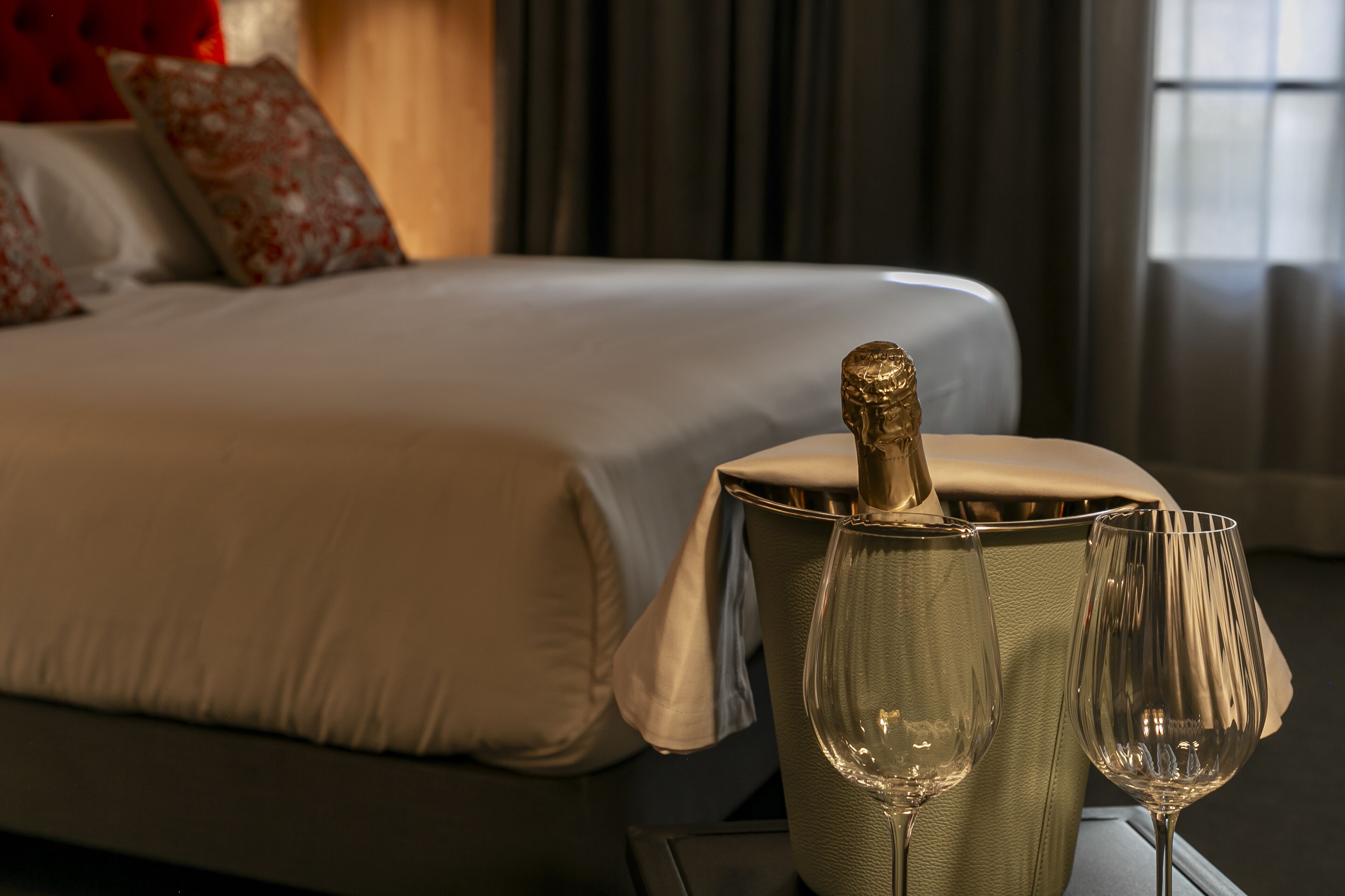 Cama com cobertura branca e almofadas florais, garrafa de champanhe em balde prateado e dois copos de vinho sobre uma mesa.