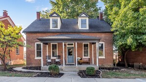 Une maison à deux étages, construite en briques, avec un porche à l’avant, deux lucarnes et un petit jardin devant, agrémenté d’un banc et de plantes en pot.