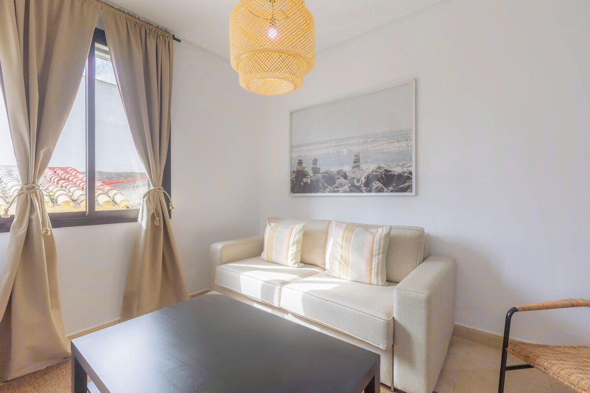 A room with a sofa, a table, a chair, a hanging light fixture, and a framed picture on the wall.