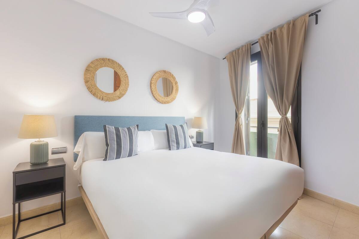A hotel room with a large bed, two bedside tables with lamps, a ceiling fan, and a window with curtains.