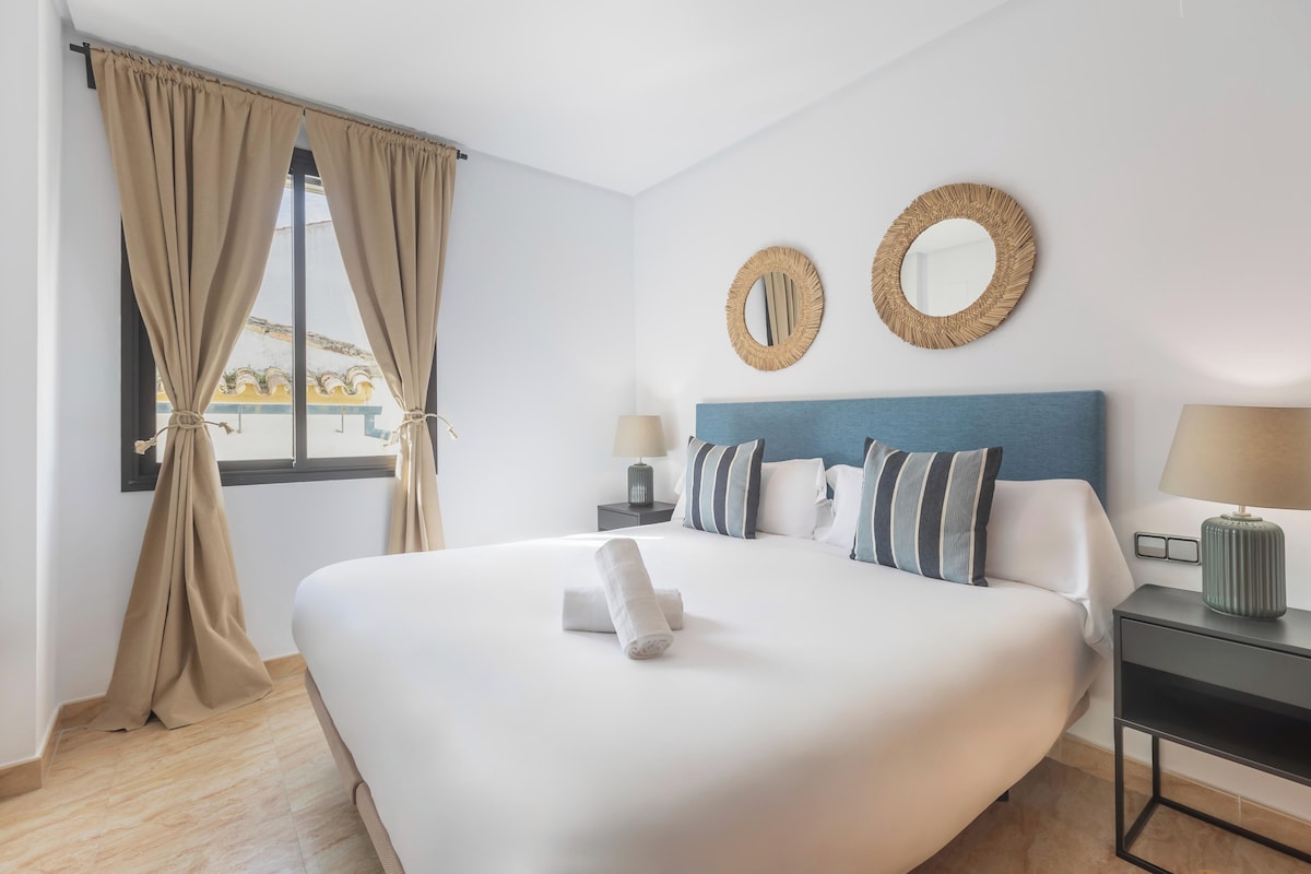 A hotel room with a large bed, two bedside tables, a window with curtains, and two wall-mounted mirrors.