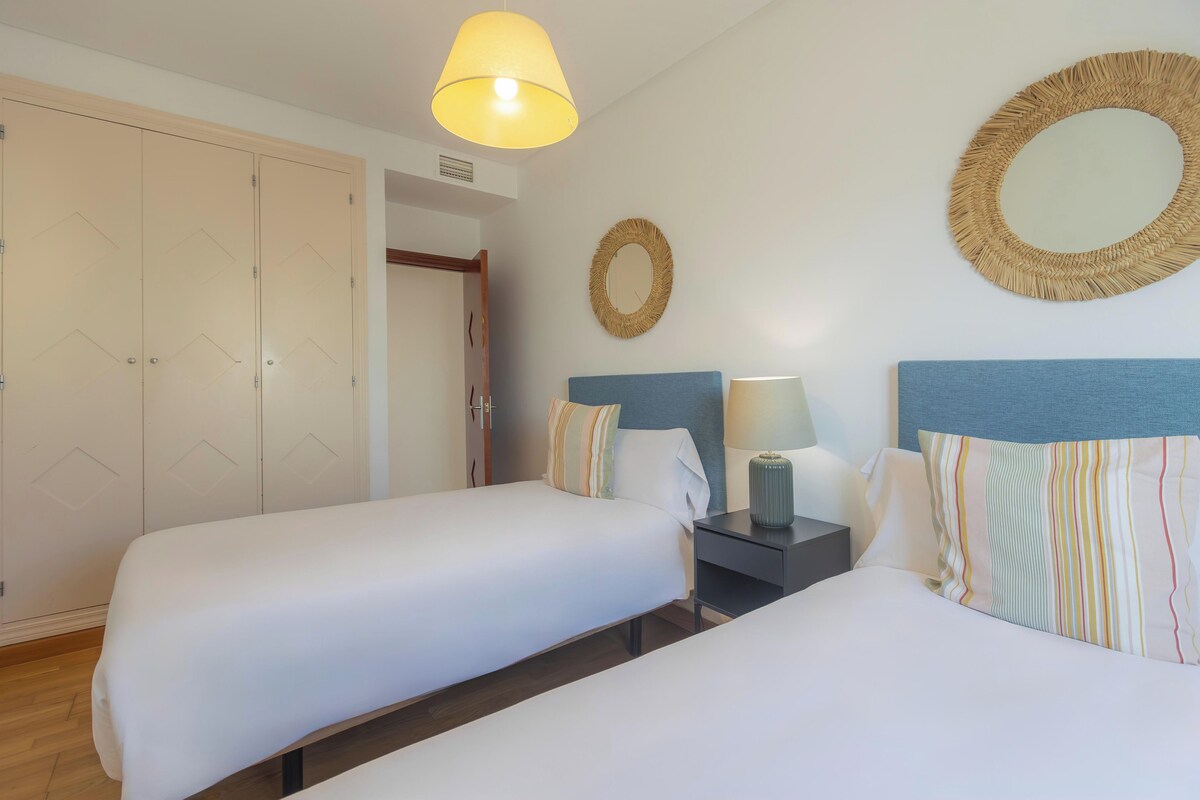 A hotel room with two beds, a nightstand, a lamp, and a mirror on the wall.