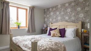 A bedroom with a bed, curtains, a window with a plant, a nightstand, and a wall with floral wallpaper. - A stone cottage that sleeps 4 guests in 2 bedrooms (Combe Martin)