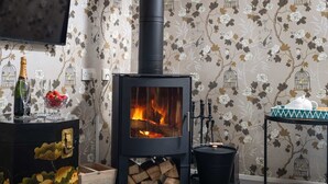 A cozy room with a wood-burning stove, a television mounted on the wall, and a decorative table with a teapot and a bowl of strawberries. - Bantam Cottage - sleeps 2 guests  in 1 bedroom (Combe Martin)