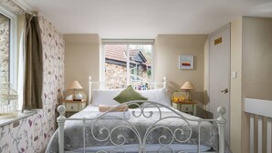 A bedroom with a white metal bed frame, floral wallpaper, a window with a view of a stone building, and two bedside tables with lamps. - Bantam Cottage - sleeps 2 guests  in 1 bedroom (Combe Martin)