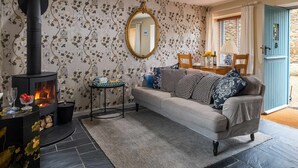 A cozy living room with a fireplace, floral wallpaper, a sofa, and a dining area. - Bantam Cottage - sleeps 2 guests  in 1 bedroom (Combe Martin)
