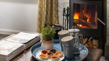 Une pièce chaleureuse avec un poêle à bois, un plateau contenant des tasses à thé et des tartes à la confiture, une plante en pot et une pile de magazines.