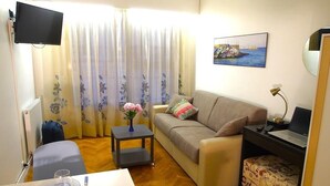 A living room with a sofa, a small table with a vase of flowers, a laptop on a desk, and a television mounted on the wall.