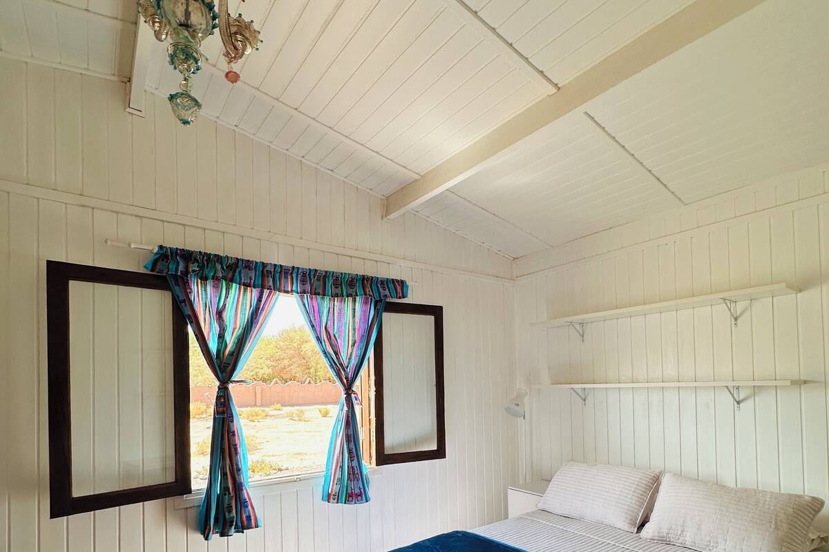 Ein Schlafzimmer mit Holzboden, einem Bett mit blauer Decke, weißen Wänden und einem Fenster mit Vorhängen.