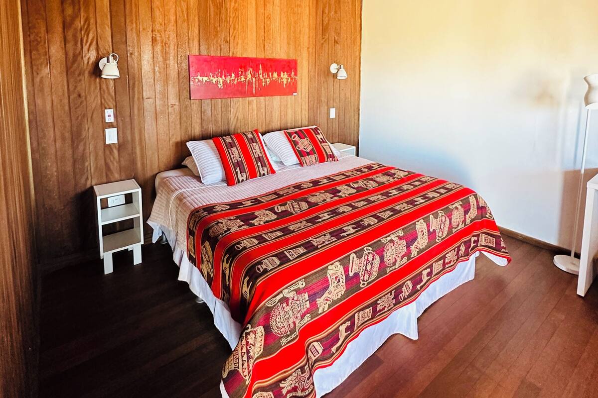 Ein Schlafzimmer mit Holzboden, einem Bett mit rotem, gemustertem Bettbezug, zwei Nachttischen und einer an der Wand befestigten Leuchte.