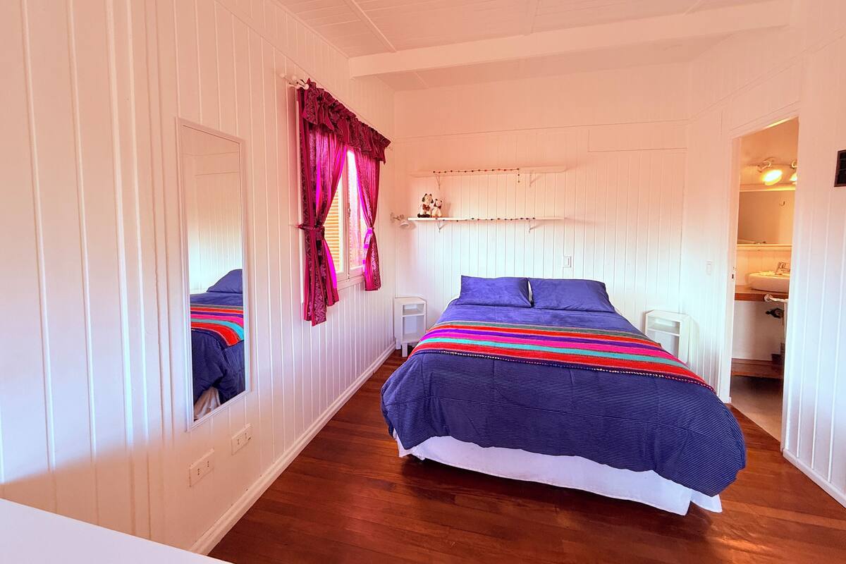 Ein Schlafzimmer mit Bett, Vorhängen am Fenster, einem Nachttisch und einem Spiegel.