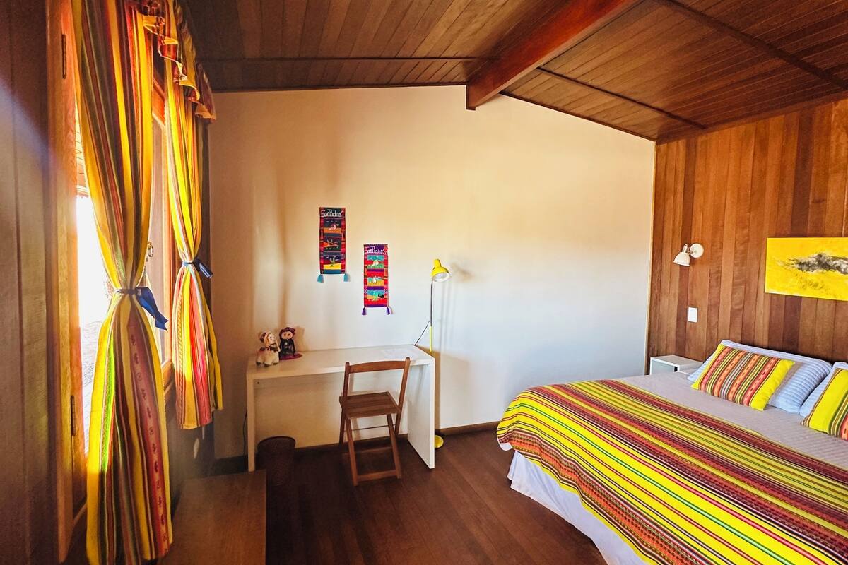 Ein Schlafzimmer mit Holzboden, einem Schreibtisch, einem Stuhl, einem Bett mit gestreifter Bettwäsche und einer Wand mit Vorhängen.